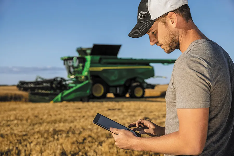 Farmer uses tablet in field; harvester behind.