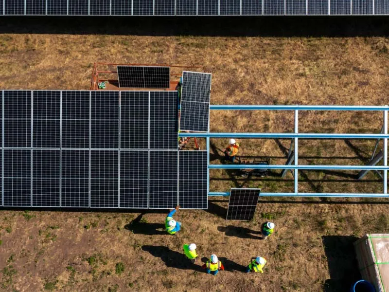 Sunlit solar farm under construction with long rows of panels on metal frames; workers in neon vests assemble the supports.
