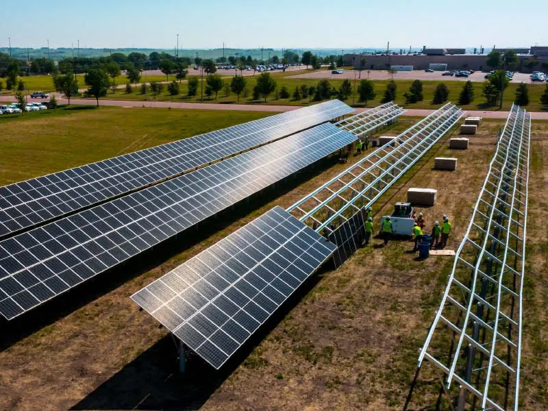 Sunlit solar farm under construction with long rows of panels on metal frames; workers in neon vests assemble the supports.