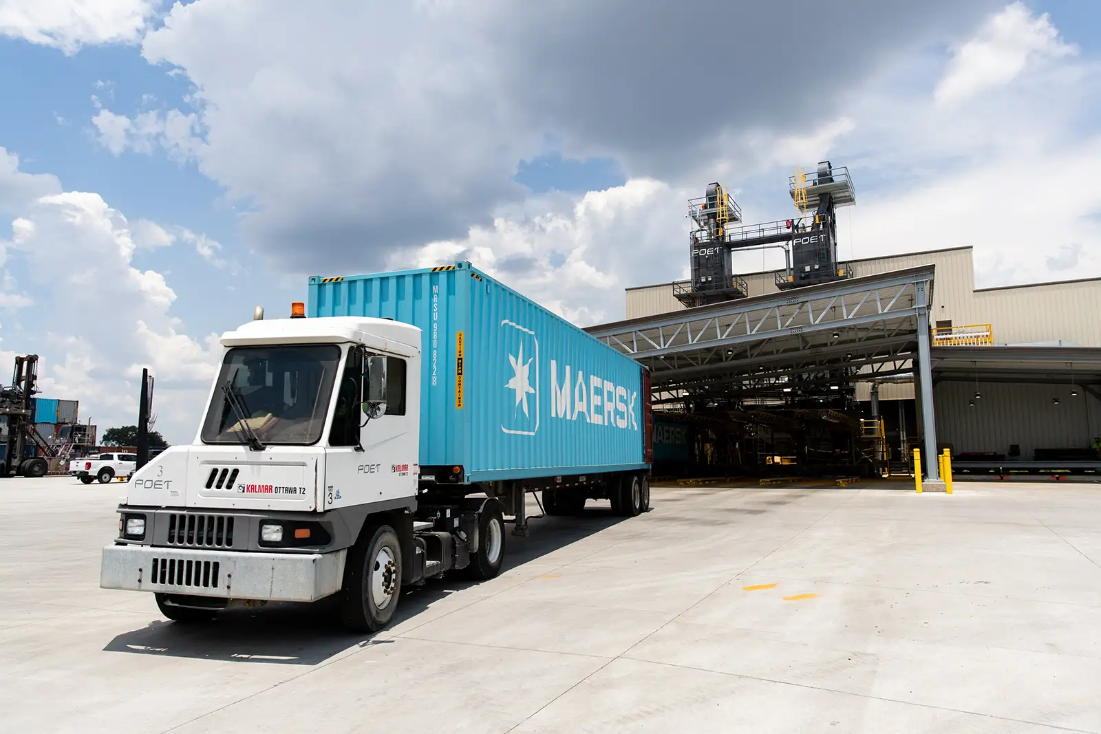 Truck hauling a blue Maersk shipping container near an industrial loading area.