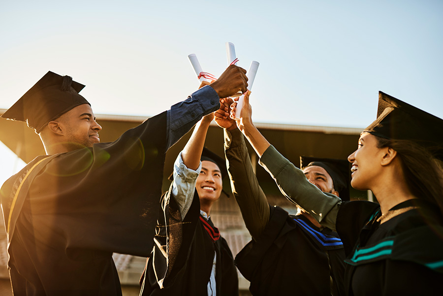 Four diverse graduates in caps and gowns raising diplomas together outdoors at sunset.
