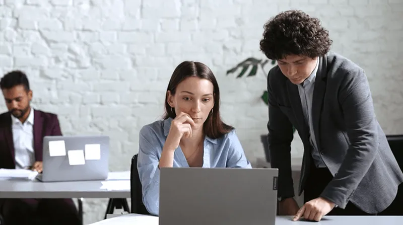 Two coworkers focused on a laptop at a desk while a third person works in the background with a laptop covered in sticky notes.