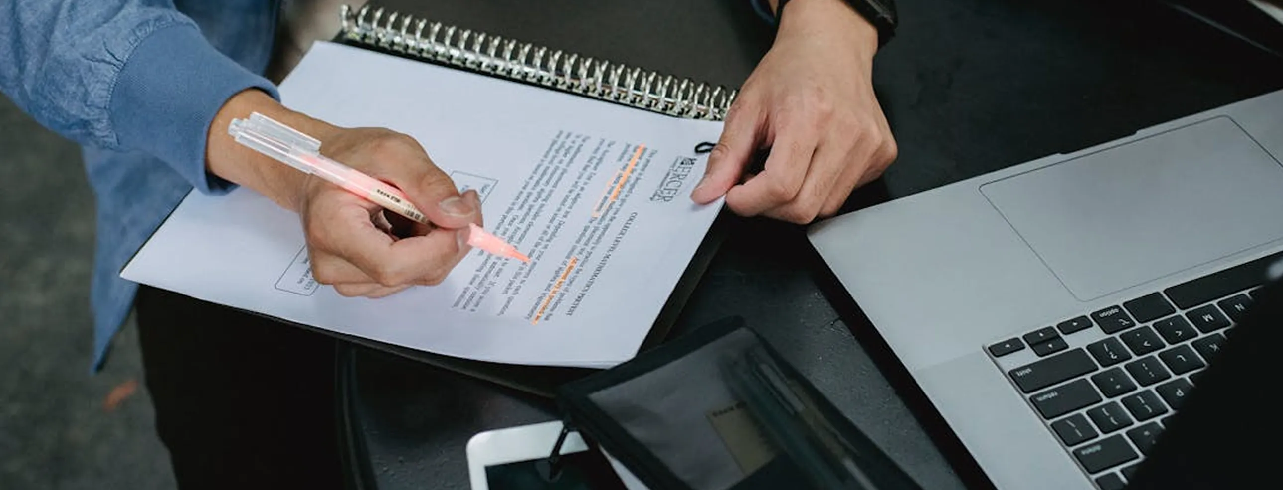 Person highlighting text on a spiral-bound document with a pink highlighter next to a laptop on a black desk.