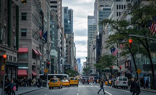 Busy city street with yellow taxis, pedestrians, and tall buildings under a cloudy sky.