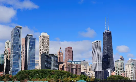 City skyline featuring various skyscrapers including a tall black building with two antennas under a blue sky with scattered clouds.