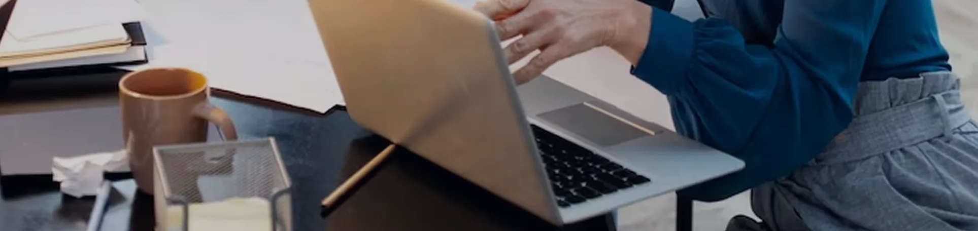 Person in blue long-sleeve shirt using a laptop on a desk with a coffee mug, pencil, and papers.