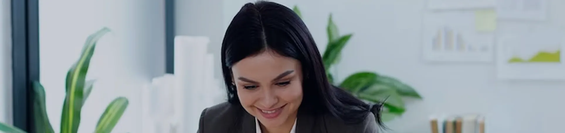 Smiling woman with dark hair working at a desk with green plants and charts in the background.