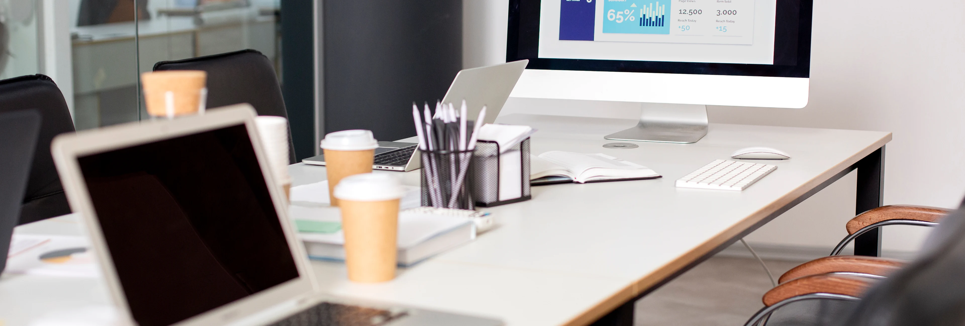 Office desk with a laptop, desktop computer displaying charts, takeaway coffee cups, notebooks, pens, keyboard, and mouse.