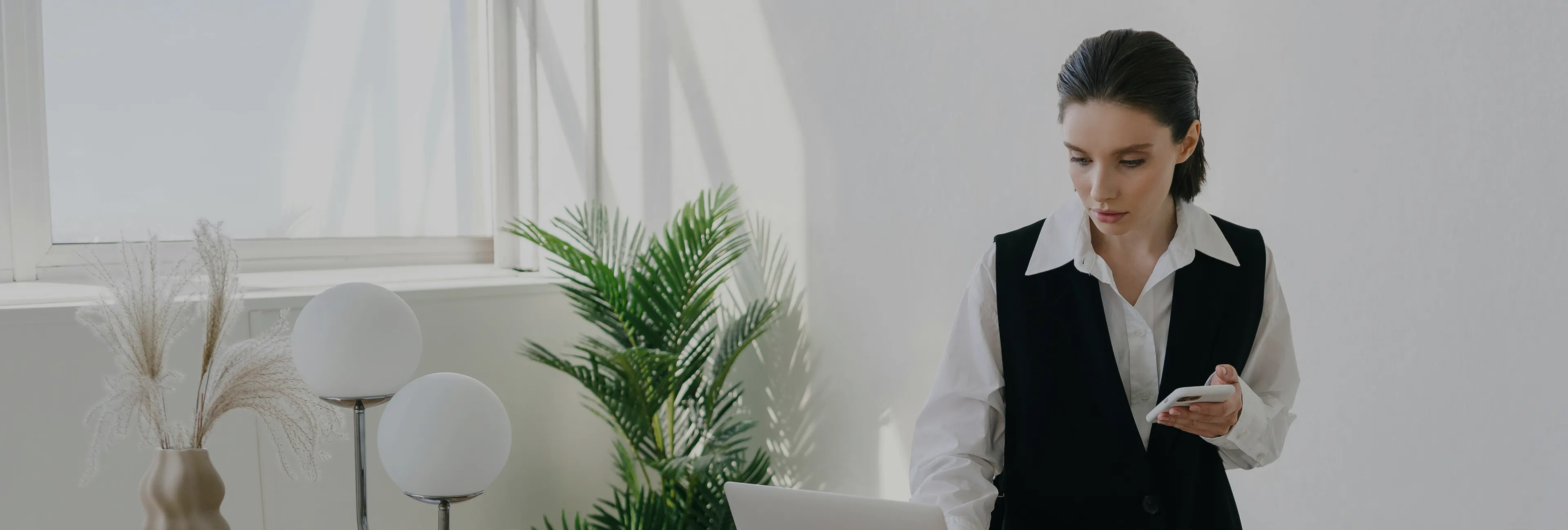 Woman in white shirt and black vest holding a smartphone and looking at a laptop in a bright room with plants and modern decor.