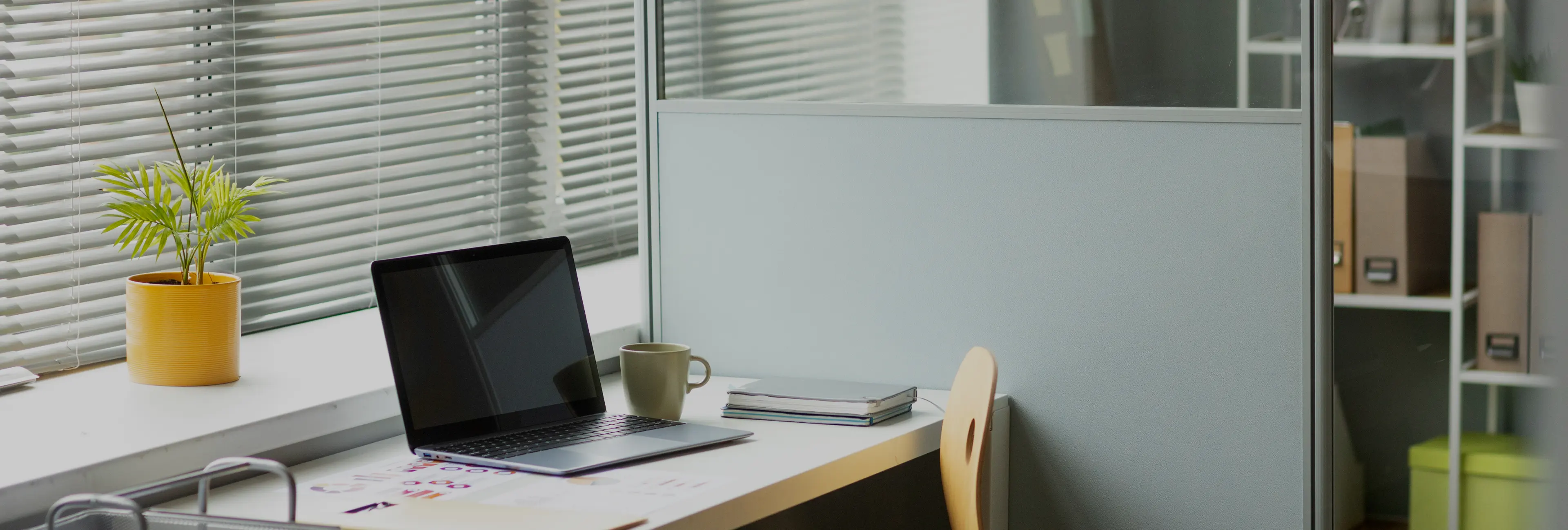 Office workspace with closed laptop, coffee mug, stacked notebooks, small potted plant, and a wooden chair next to a gray cubicle wall.