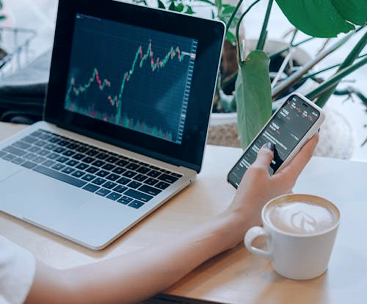 Person holding a smartphone showing financial data next to a laptop displaying a stock market chart, with a cup of coffee and green plants nearby.