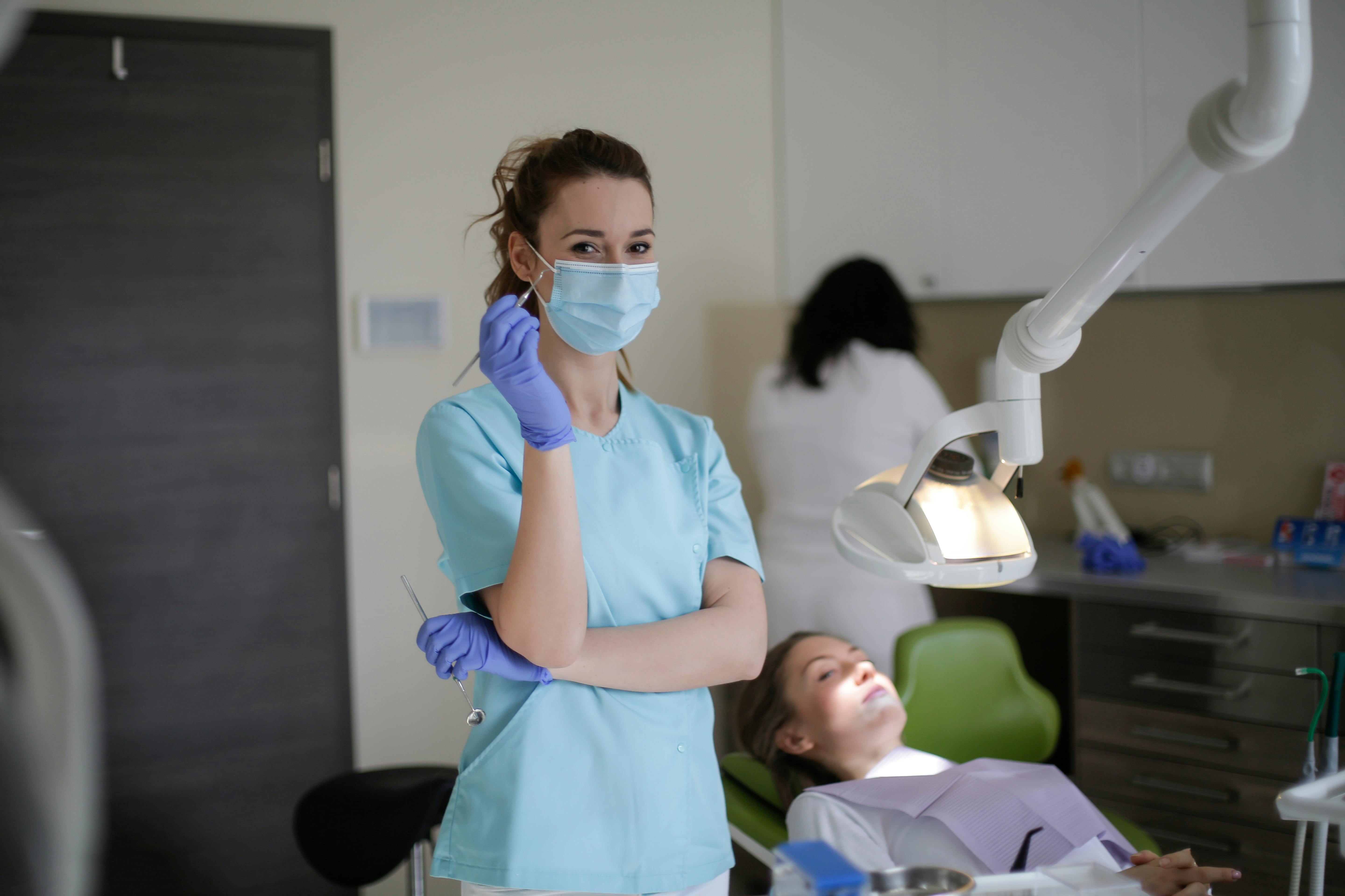 Female dentist wearing a mask and gloves holding dental tools in a clinic with a patient reclining in the dental chair.