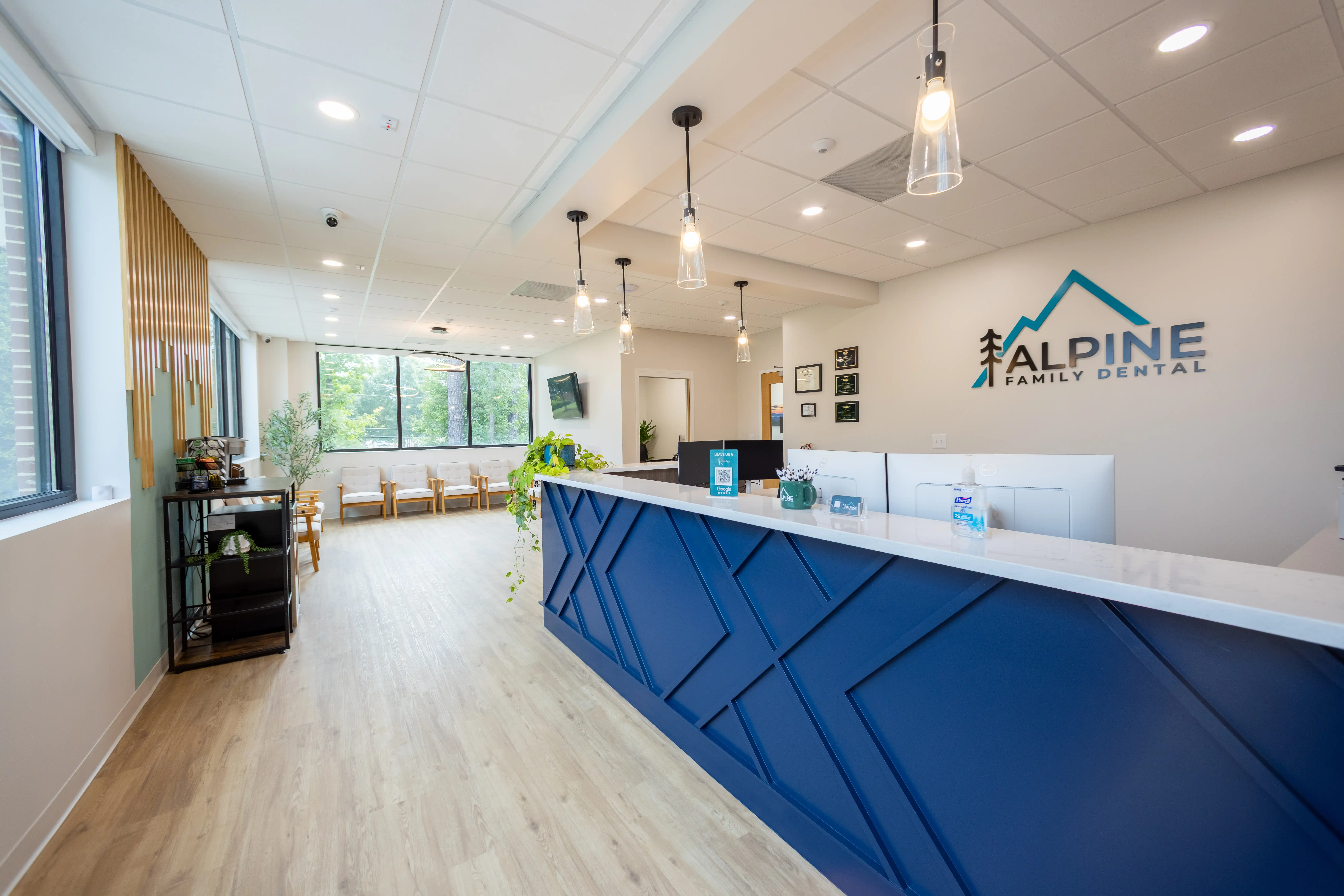 Modern dental office reception area with a blue front desk, hanging pendant lights, seating area with chairs, and large windows showing greenery outside.