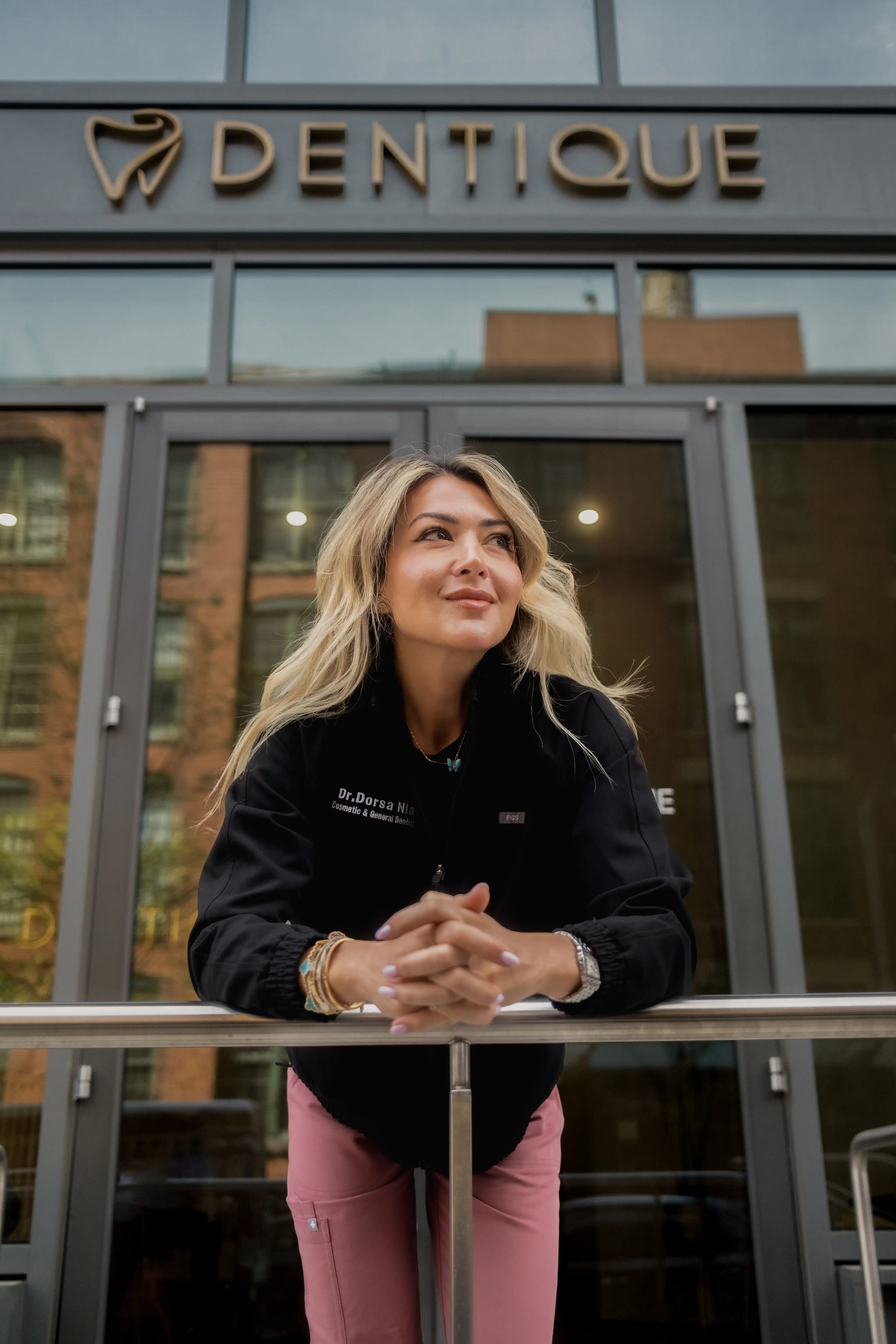 Blonde woman in a black jacket and pink pants leaning on a railing outside a building with a Dentique sign.
