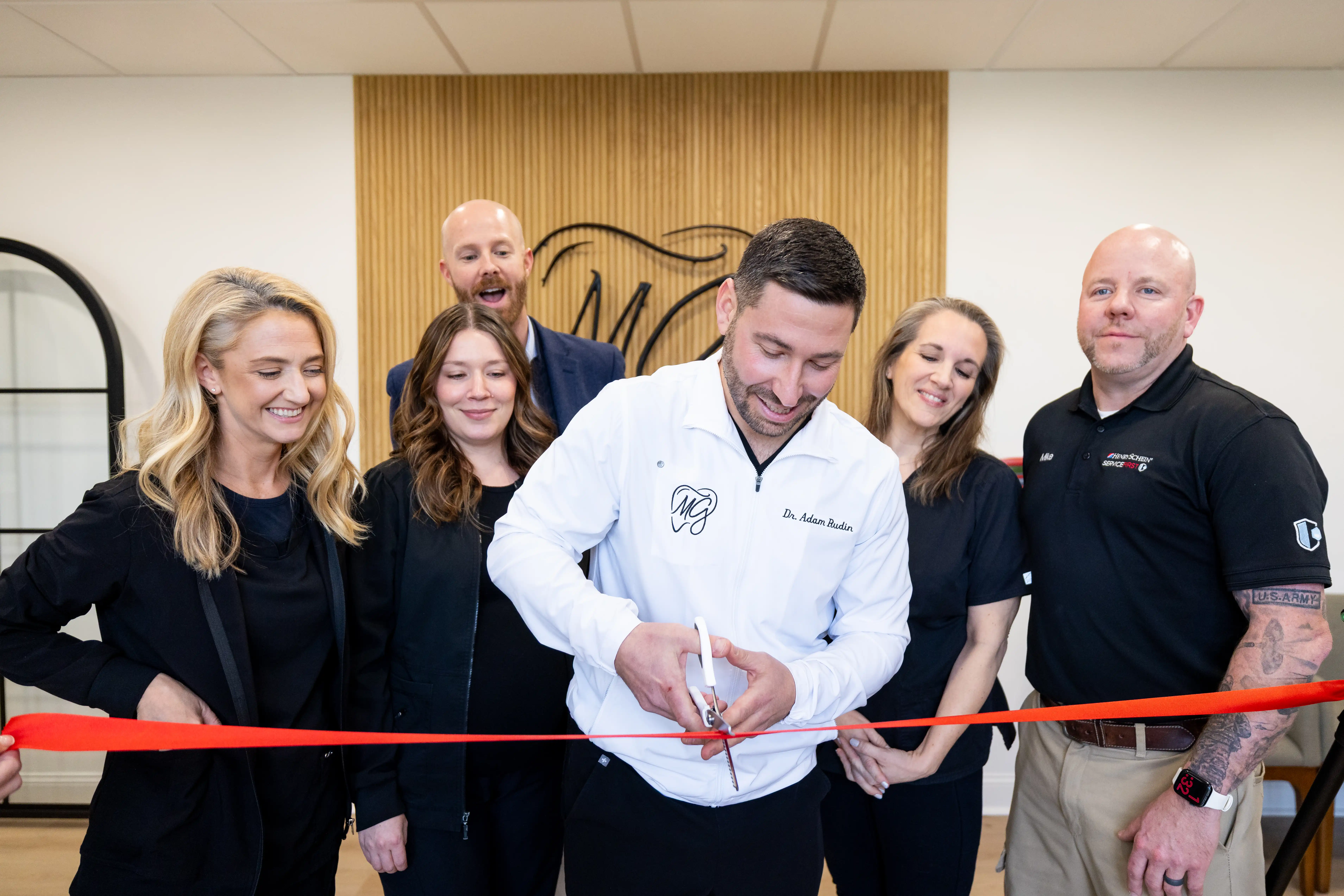 Man in white jacket cutting a red ribbon at a ceremony with five people smiling and standing behind him.