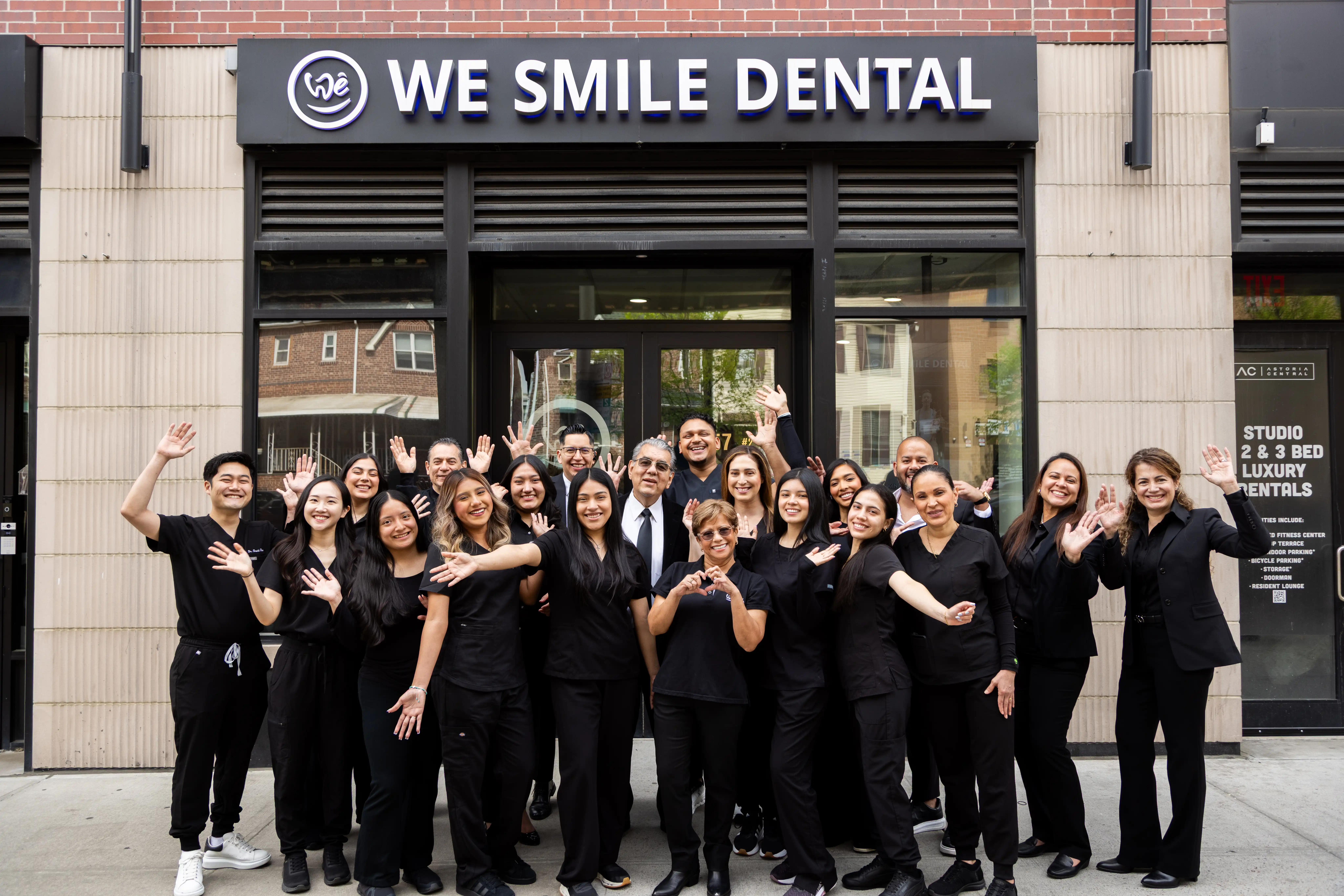 Group photo of smiling dental staff in black uniforms waving in front of We Smile Dental clinic.