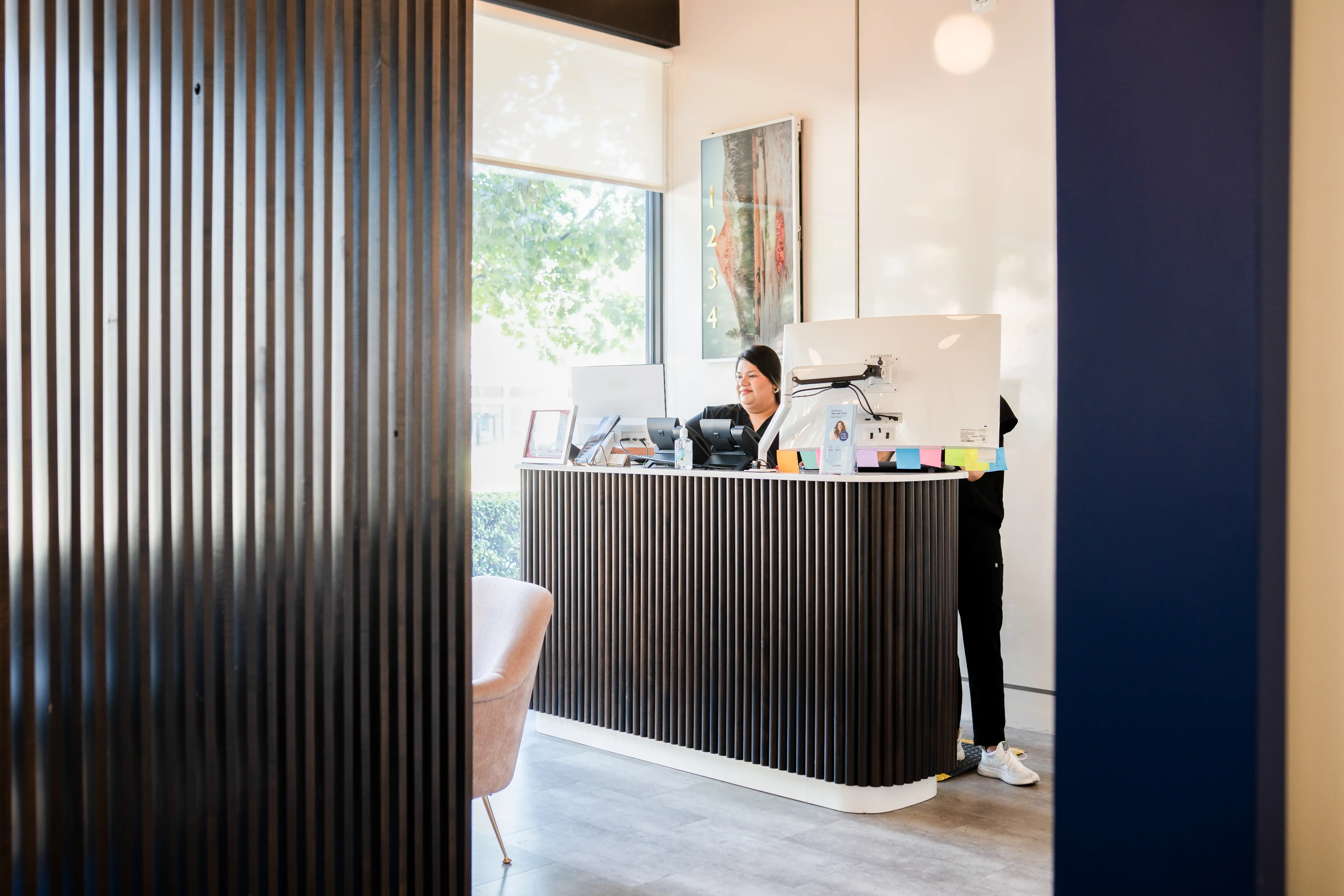 Reception area with two people behind a modern dark wood reception desk and a window showing greenery outside.