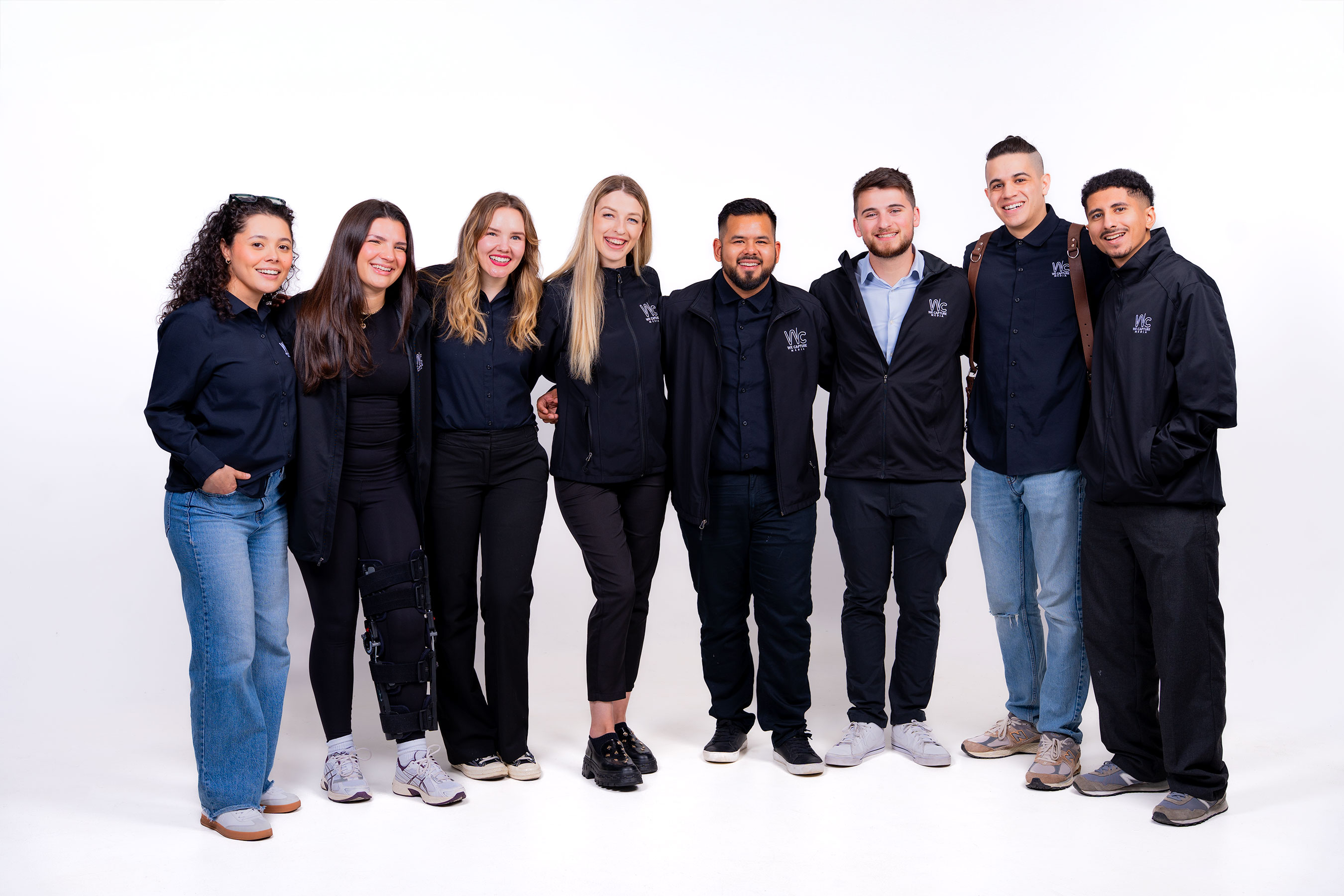 Group of eight WeCapture Dental team members  side by side smiling, wearing black jackets or shirts with the logo 'WC' ready for dental marketing. 
