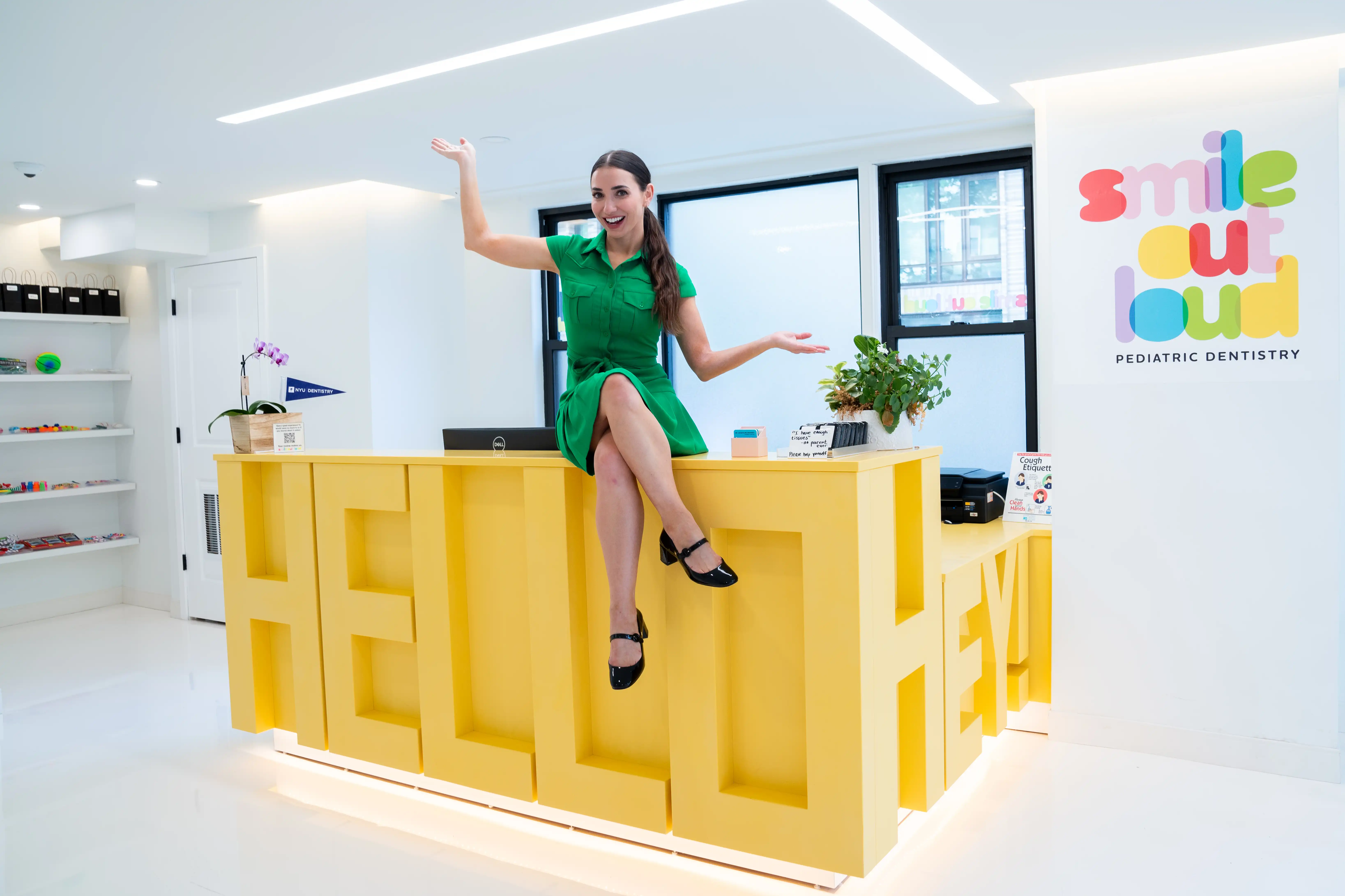 Dr. Jacquline Dikansky in a green dress sitting on a yellow reception desk with large letters spelling HELLO and HEY in a bright pediatric dentistry office.