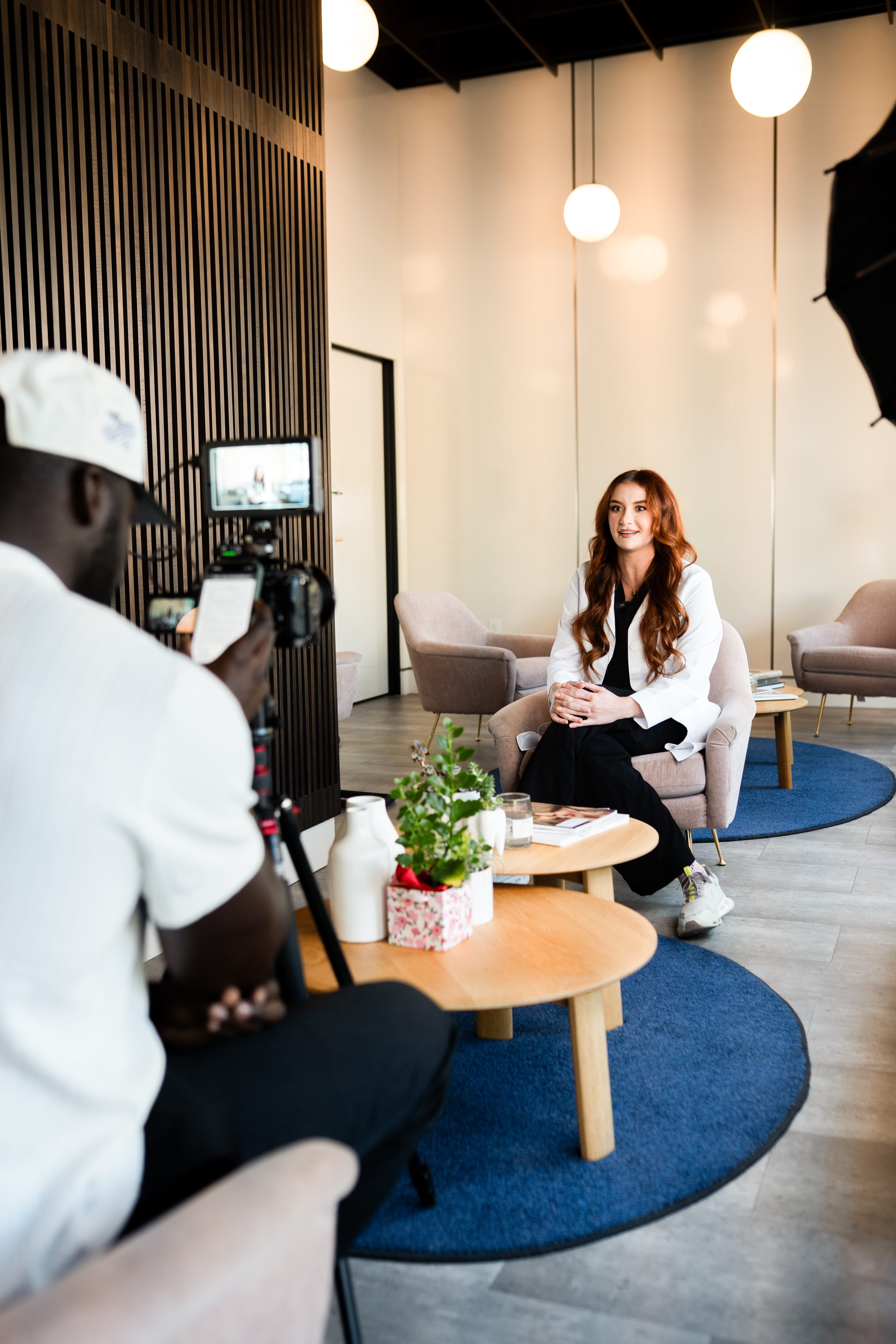 Female professional seated in an armchair being recorded on camera in a well-lit, modern office lounge.