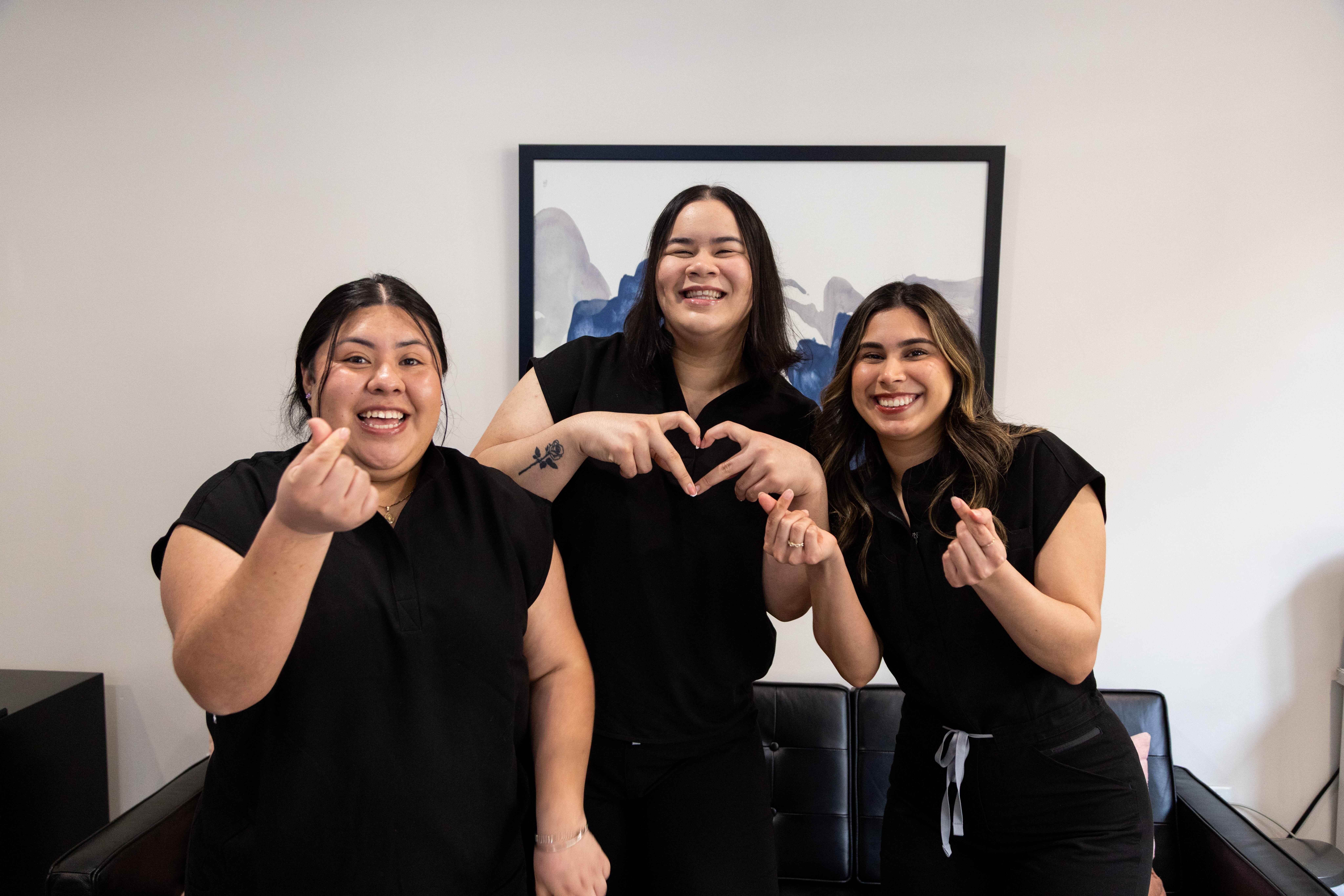 Three smiling women in black uniforms standing indoors, making heart and finger heart gestures.