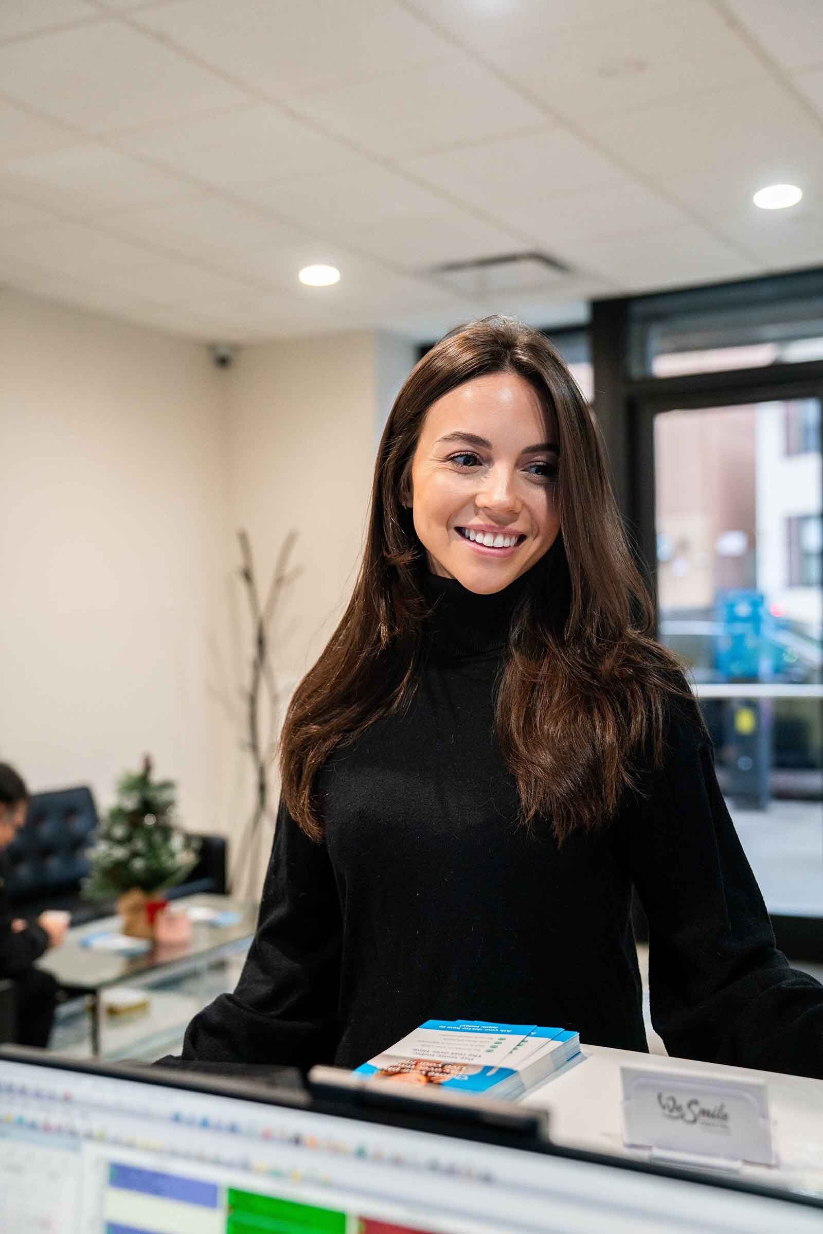 Smiling woman with long brown hair wearing a black turtleneck sweater standing in a modern office reception area.