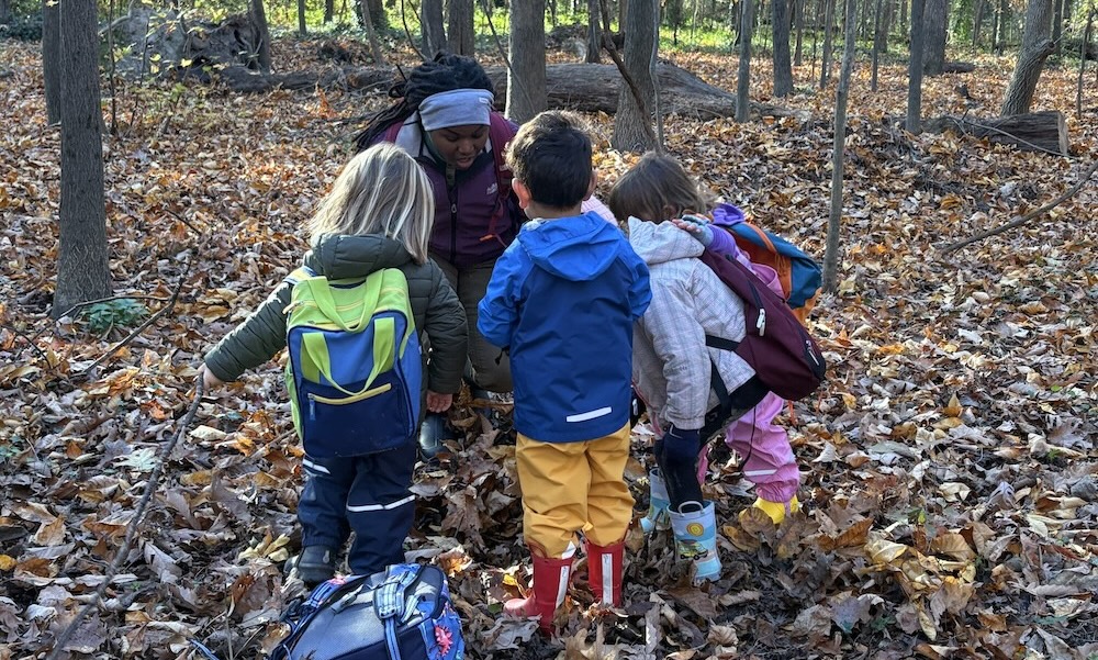 Group of children with backpacks and colorful rain boots gathered around an adult in a wooded area covered with fallen leaves.
