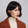 Portrait of a woman with short black hair wearing a white turtleneck and brown blazer against a neutral background.
