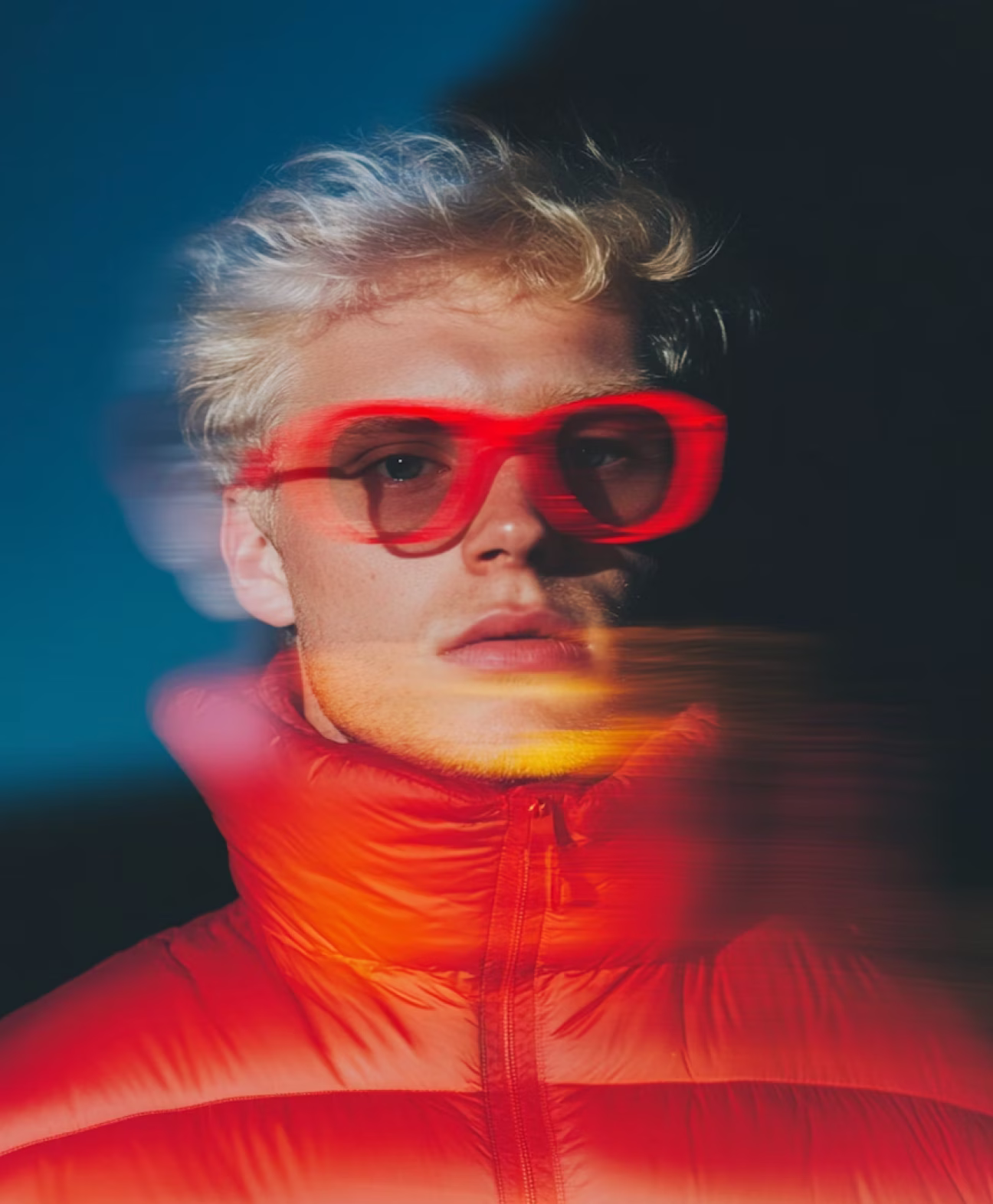 Young man with blonde hair wearing red tinted glasses and a red puffer jacket against a dark background with motion blur effects.