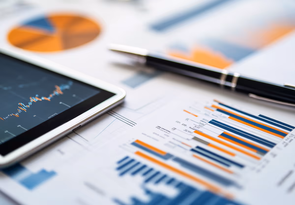 Close-up of financial documents with bar and pie charts, a tablet displaying a line graph, and a pen on a desk.