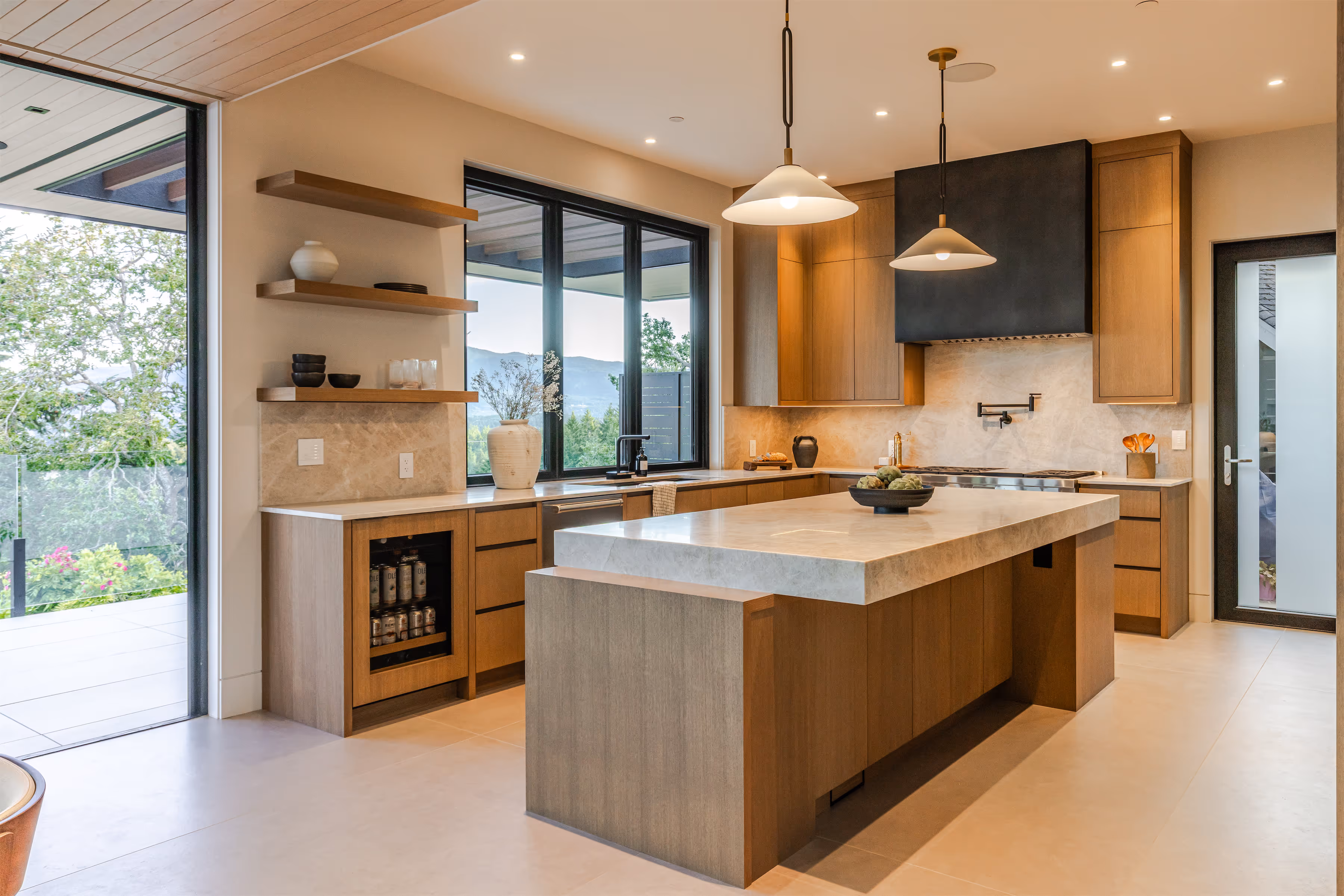 Modern kitchen with wooden cabinets, marble island countertop, pendant lights, and large window overlooking greenery.