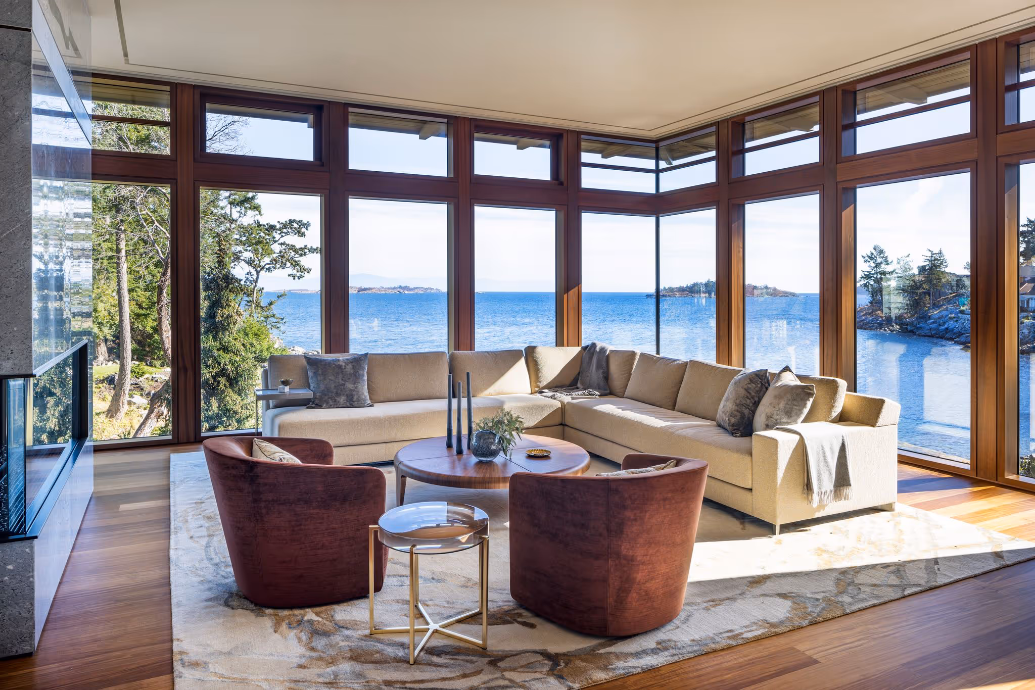 Modern living room with beige sectional sofa, two burgundy armchairs, wooden coffee table, and large windows overlooking a scenic ocean view.