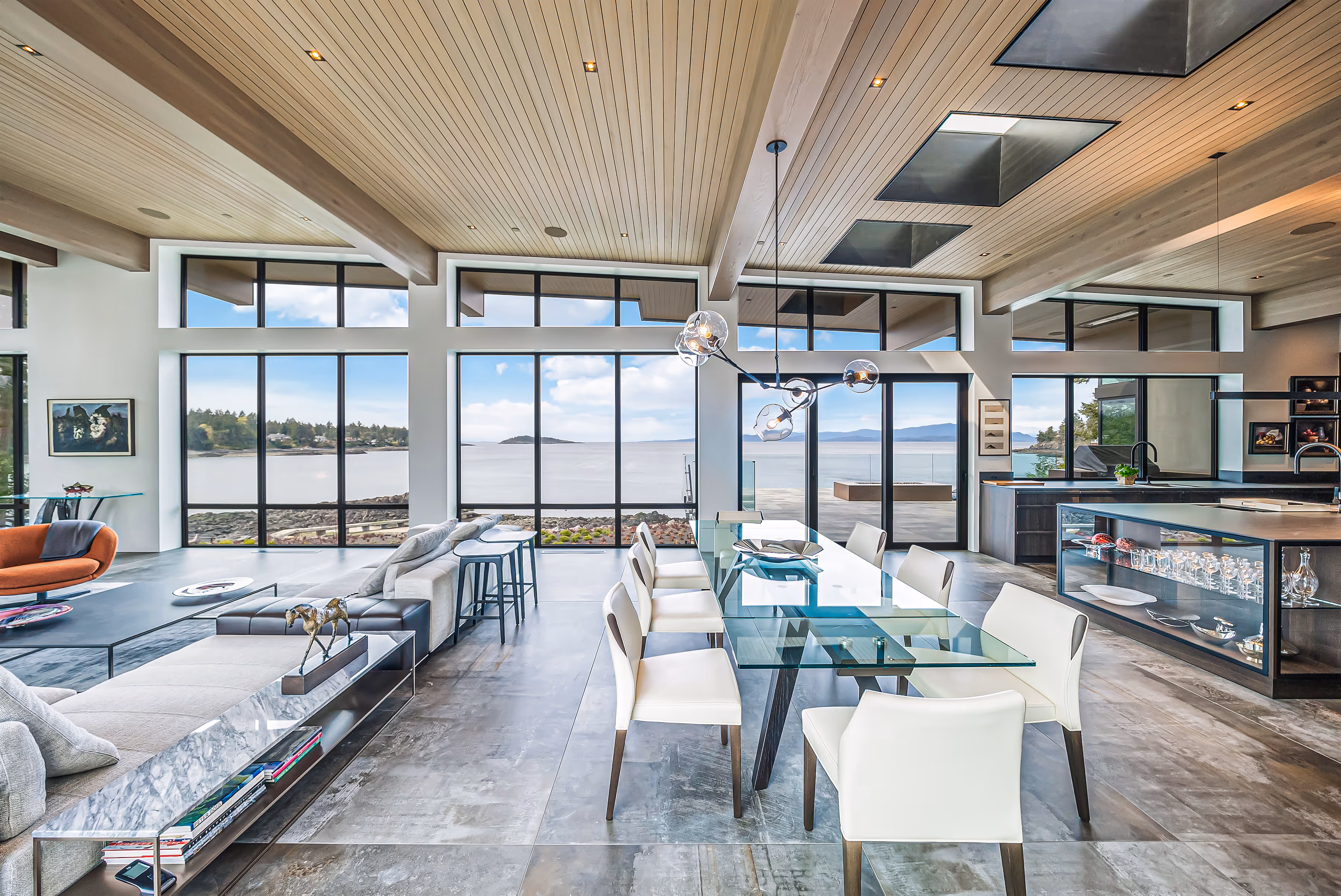 Modern open-concept living and dining area with glass dining table, white chairs, large sectional sofa, and floor-to-ceiling windows overlooking a waterfront view.
