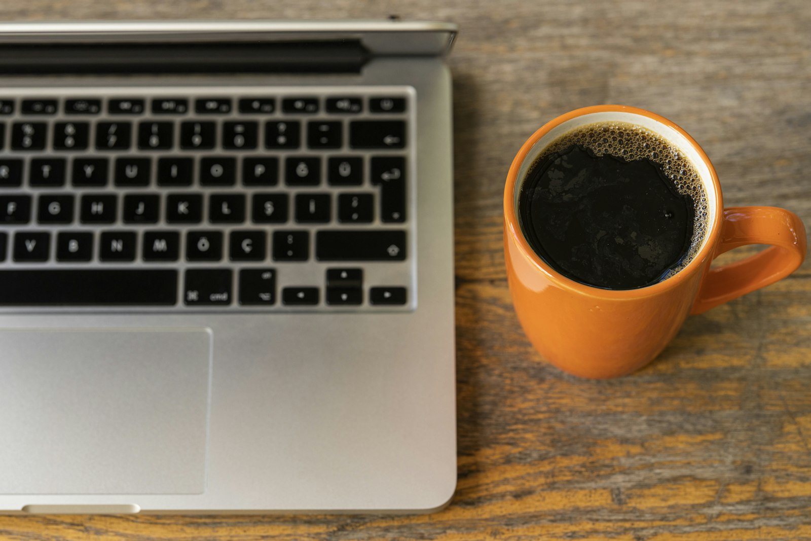 Black coffee in an orange mug beside a laptop keyboard on a wooden table