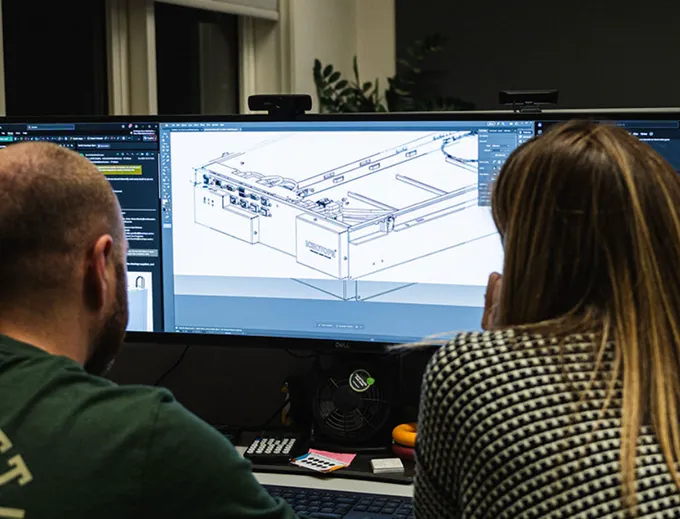 Two people working together at a computer displaying a detailed technical drawing or CAD model.