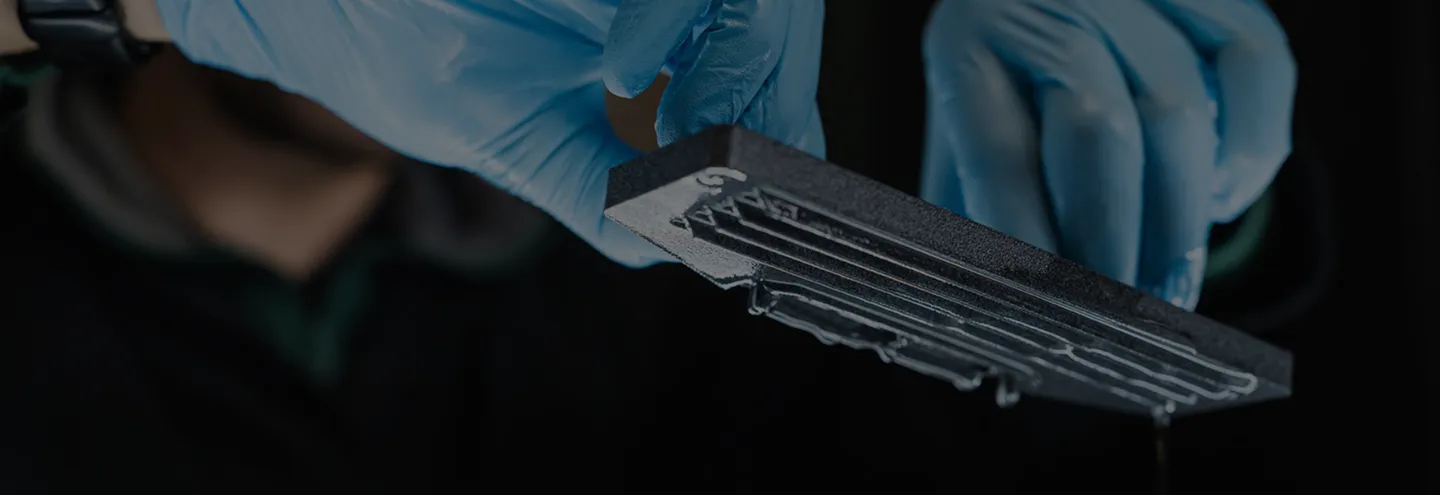 Hands wearing blue gloves holding a black rectangular object with clear liquid dripping from it against a dark background.