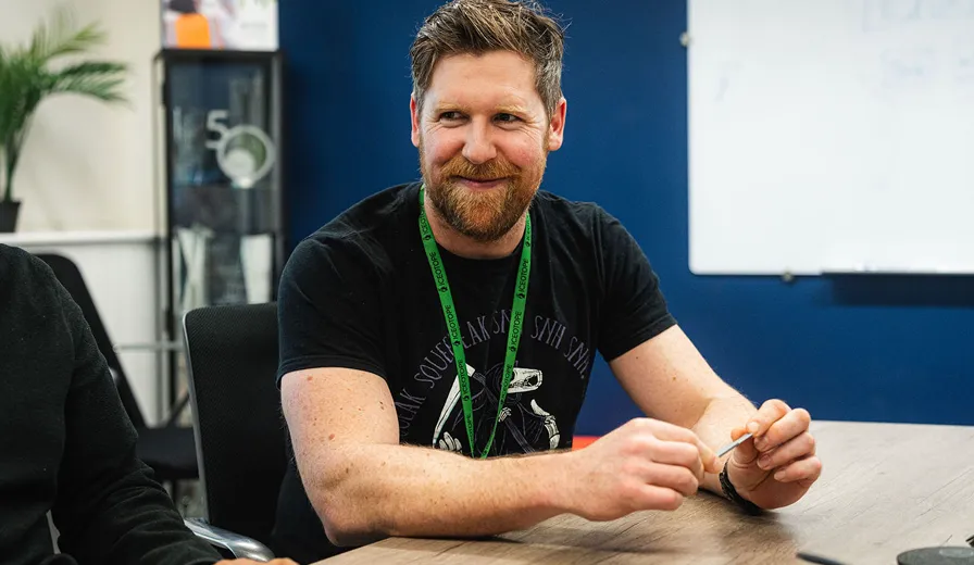 Man with a beard wearing a black t-shirt and green lanyard smiling while sitting at a desk in an office.