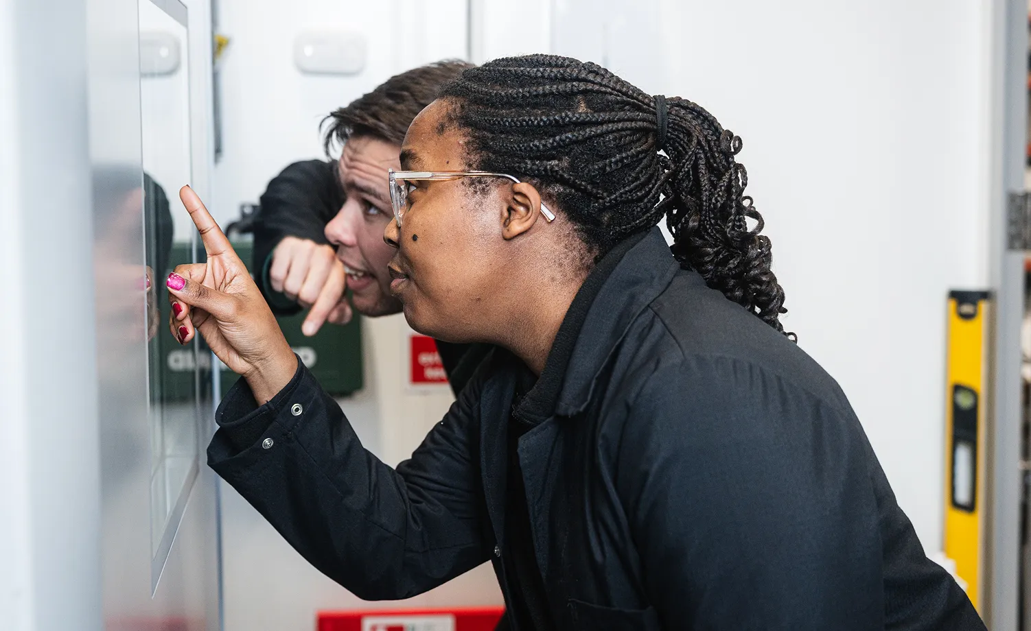 Two people closely examining a wall-mounted control panel, one pointing at it while the other watches attentively.