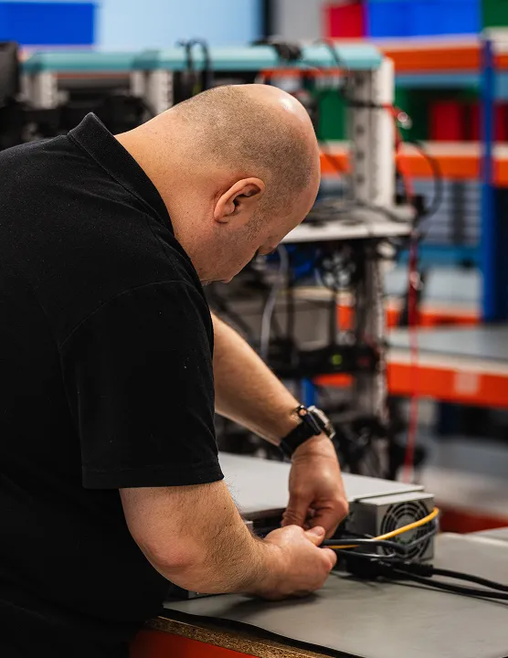 Man with a black shirt working on electronic equipment with cables on a table in a workshop.