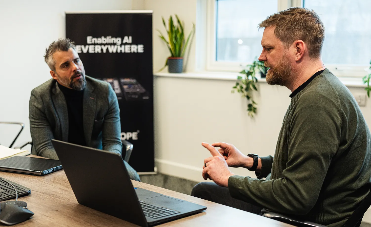 Two men having a discussion in an office with a laptop on the table and a banner in the background that reads 'Enabling AI EVERYWHERE'.