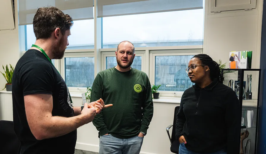 Three people engaged in a discussion in a bright office room with windows and plants.