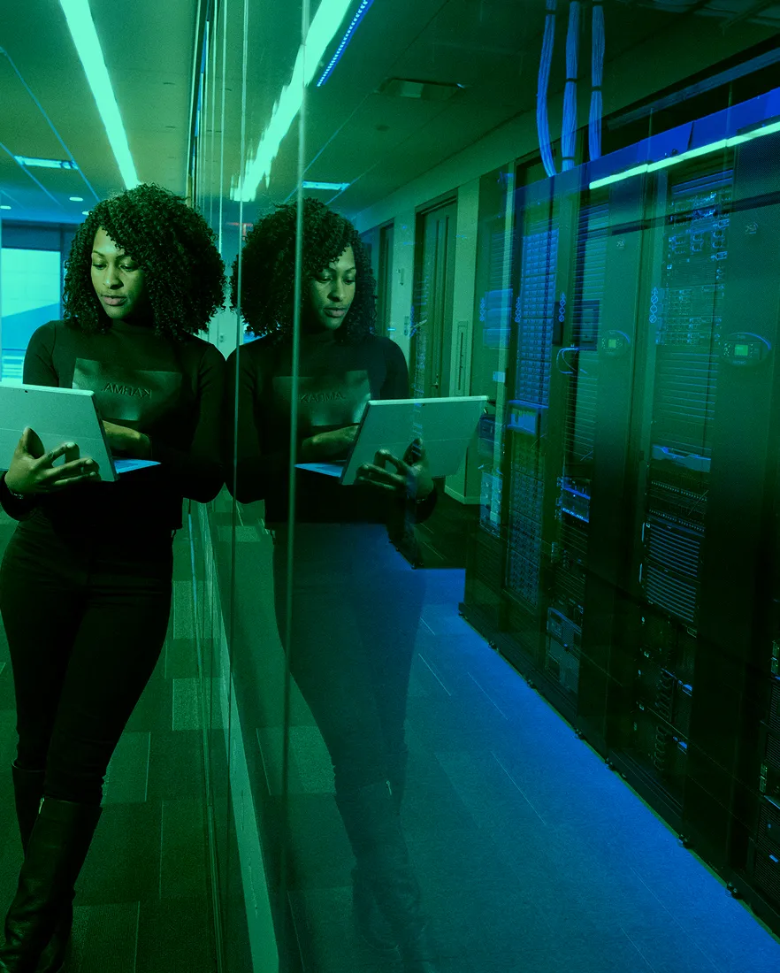 Woman in black holding a tablet standing next to a glass wall reflecting server racks in a data center.