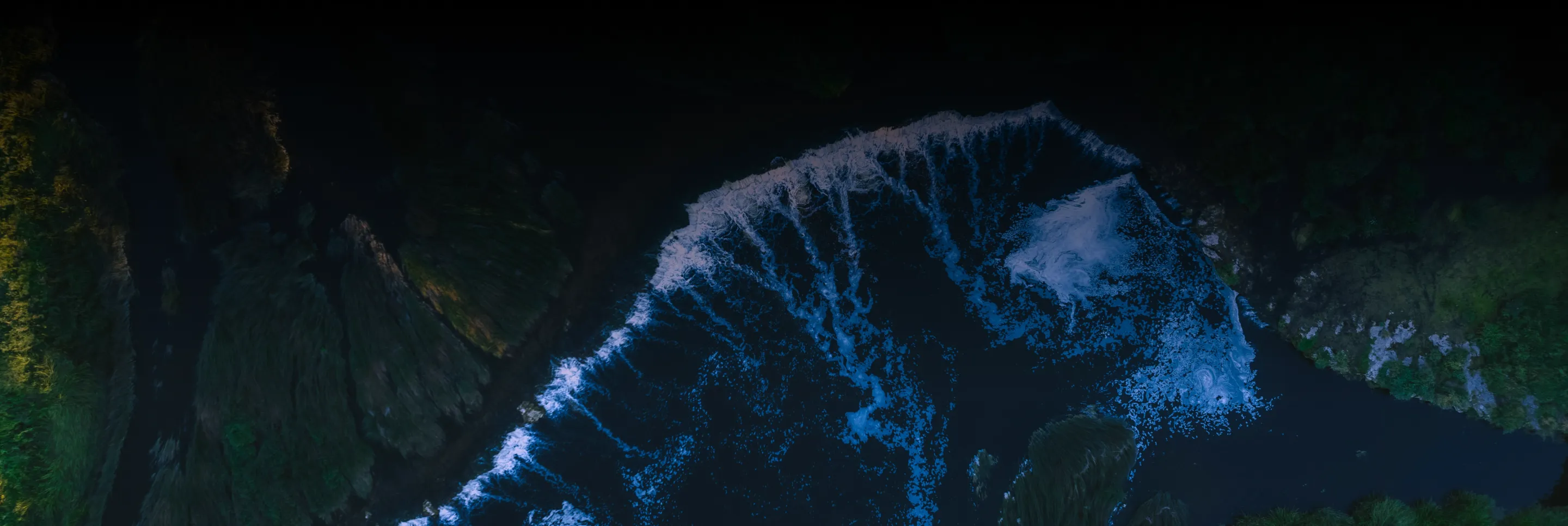 Aerial view of foamy sea waves crashing against dark rocky shores and green vegetation.