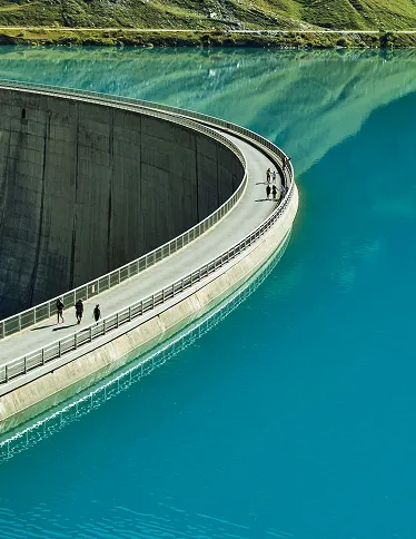 People walking along the curved edge of a large dam with turquoise water on one side and green mountains reflected in the water.