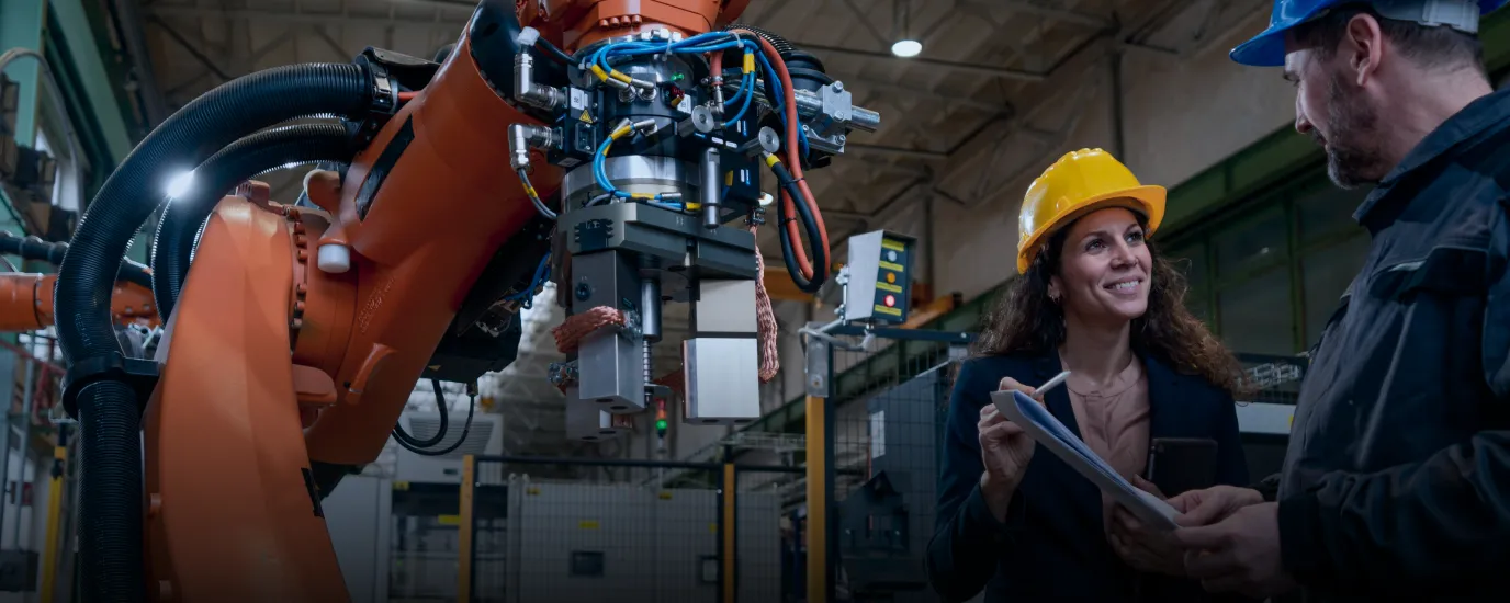 Two industrial workers with helmets in a factory next to a robotic arm and machinery.