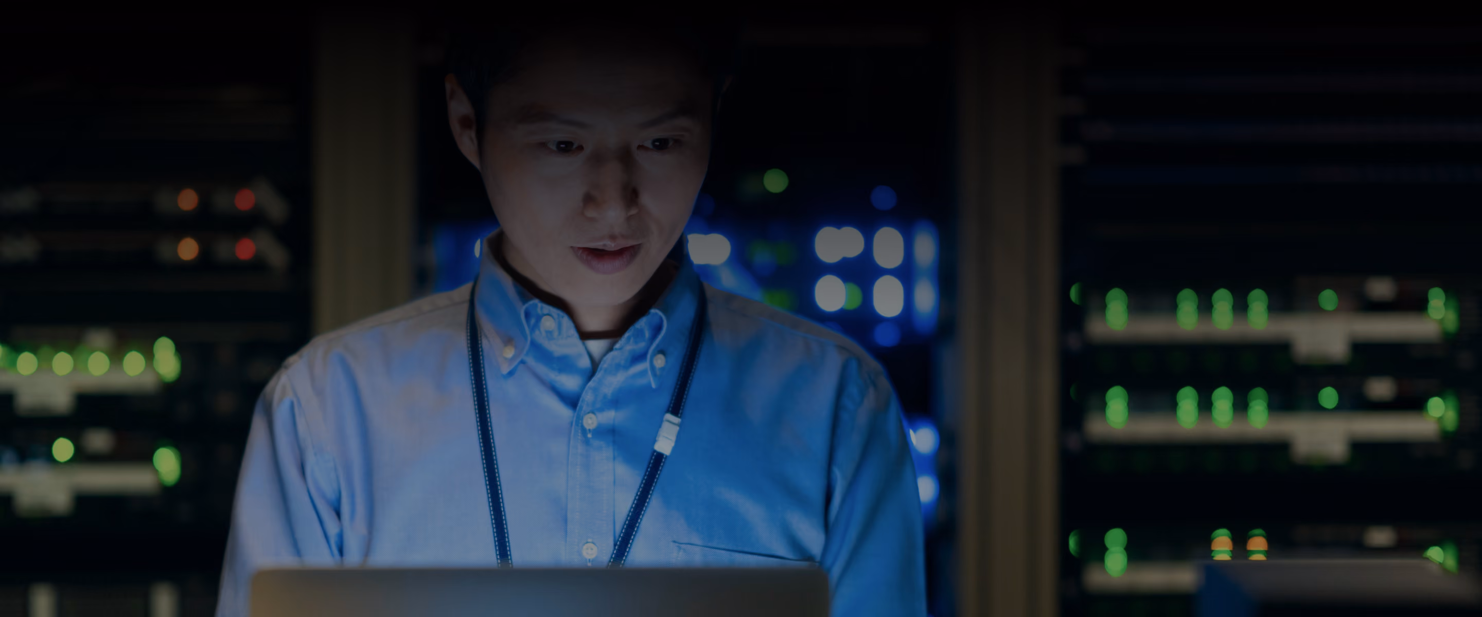 Man in blue shirt working on a laptop with blurred server racks and green lights in the background.
