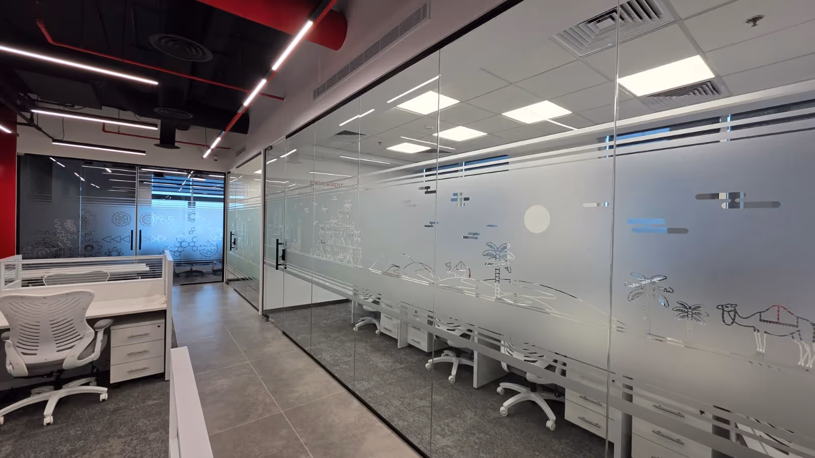 Modern office interior with glass-walled meeting rooms featuring frosted designs of landscapes and gears, white office chairs, and gray tiled floors.