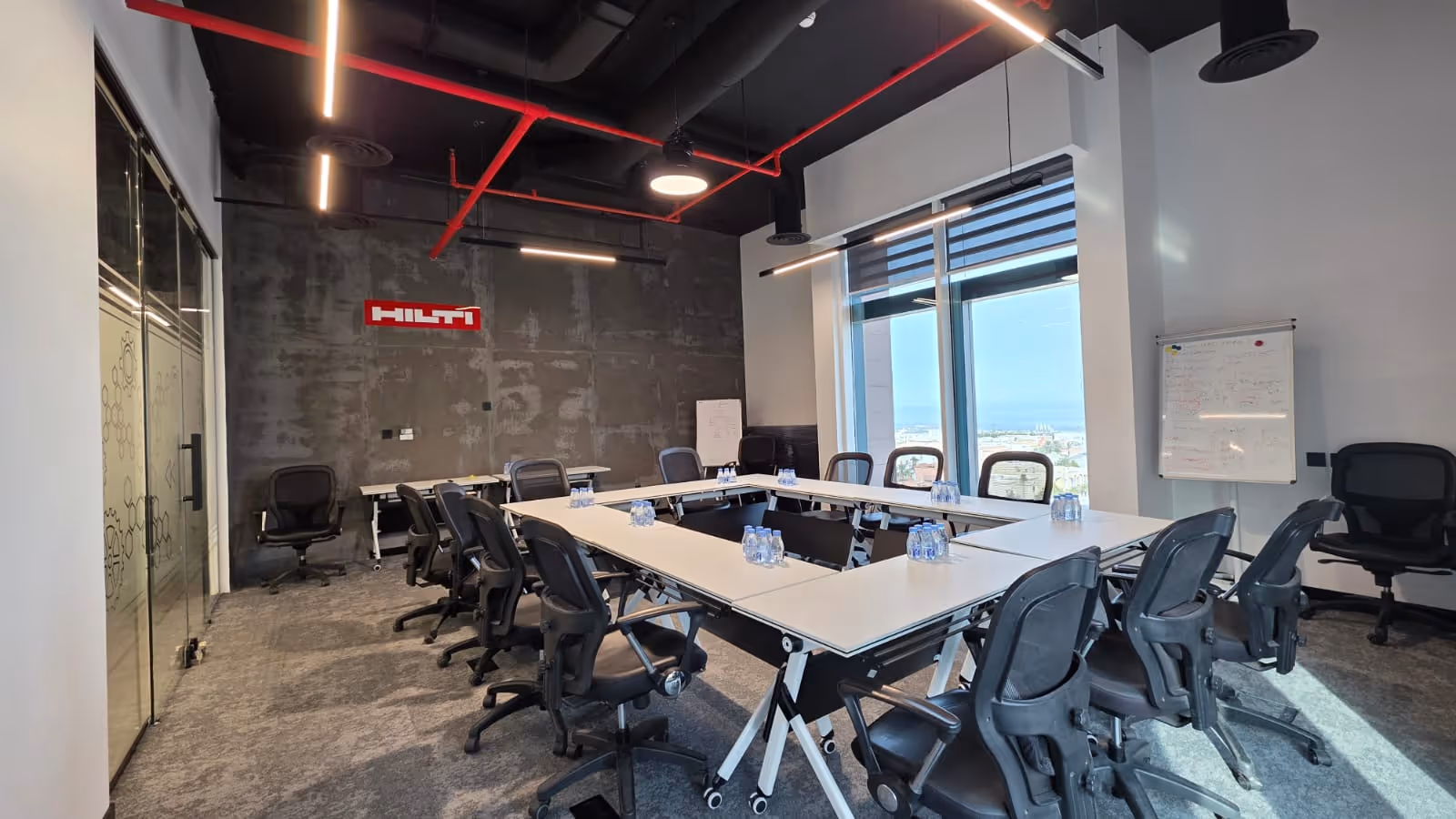 Modern conference room with U-shaped tables, black mesh chairs, water bottles on tables, large windows, and Hilti logo on concrete wall.