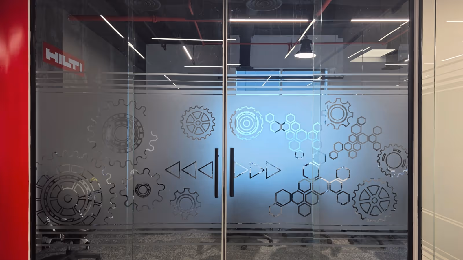 Glass office doors with etched gear and hexagonal patterns, featuring black handles and a red wall with the Hilti logo on the left.