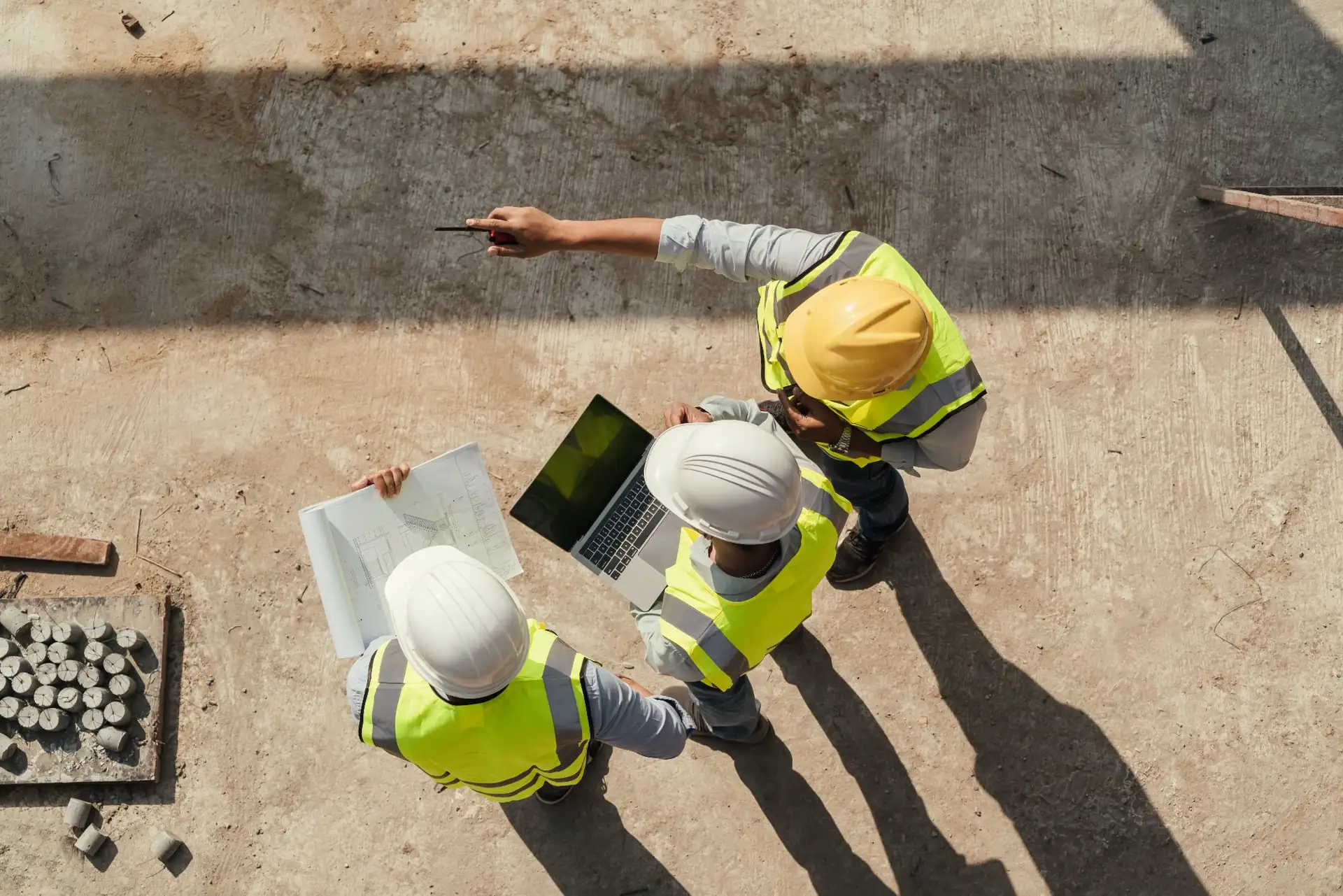 Three construction workers wearing hard hats and reflective vests reviewing blueprints and a laptop at a construction site.